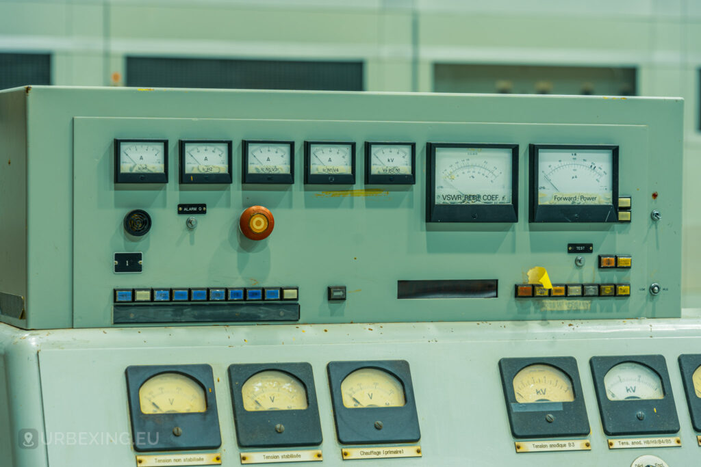 A close-up view of an old control panel at "Lost Frequencies," an abandoned radio transmitting facility. The panel features various dials, gauges, and switches, including a prominent red emergency stop button. This detailed shot highlights the intricate components that make it a fascinating subject for urban exploring.