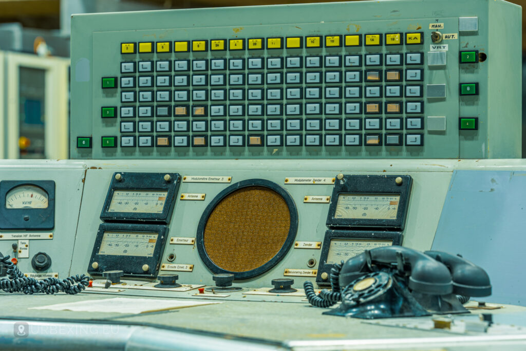 A detailed shot of a control console in the "Lost Frequencies" abandoned radio transmitting facility. The image showcases numerous buttons, switches, and a central speaker, with vintage telephones nearby. This scene is a prime example of urban exploring, revealing the technical setup of past operations.