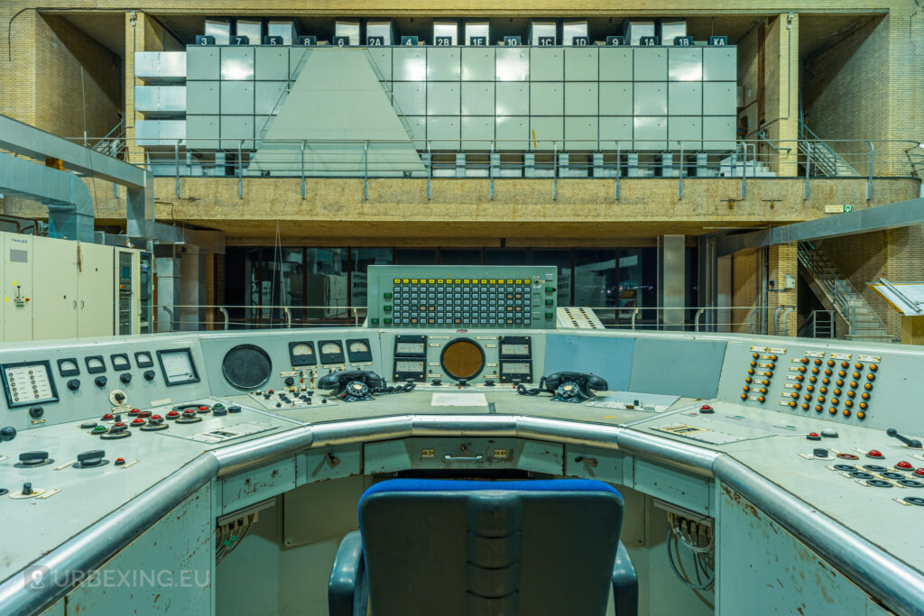 A comprehensive view of the control room in "Lost Frequencies," an abandoned radio transmitting facility. The central control desk is surrounded by various panels, switches, and telephones, with a large wall of control units in the background. This image captures the essence of urban exploring in a historic technological site.