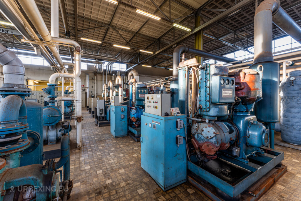 A room inside the abandoned EMGO factory in Lommel, Belgium, filled with rows of blue Atlas Copco industrial machines and a network of pipes, showcasing the extensive and decayed industrial infrastructure.