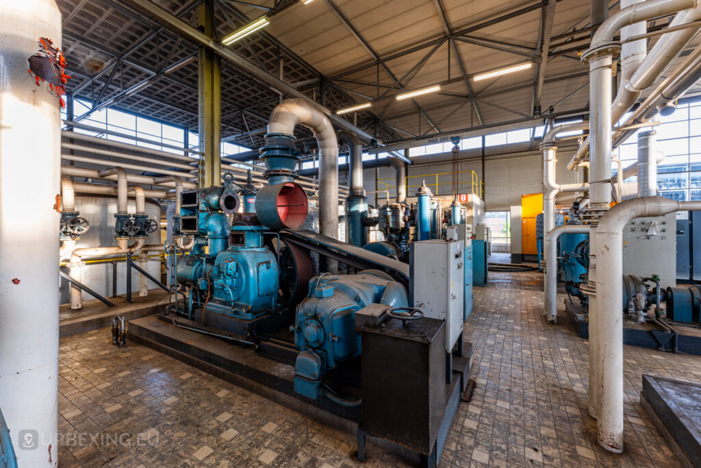 An extensive industrial area inside the abandoned EMGO factory in Lommel, Belgium, filled with large blue Atlas Copco machines and a complex network of pipes, highlighting the decayed state of the industrial setup.