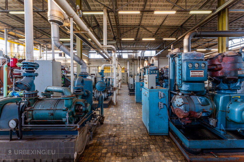 An area inside the abandoned EMGO factory in Lommel, Belgium, featuring rows of large blue Atlas Copco machines and a complex network of pipes, showcasing the extensive and decayed industrial infrastructure.