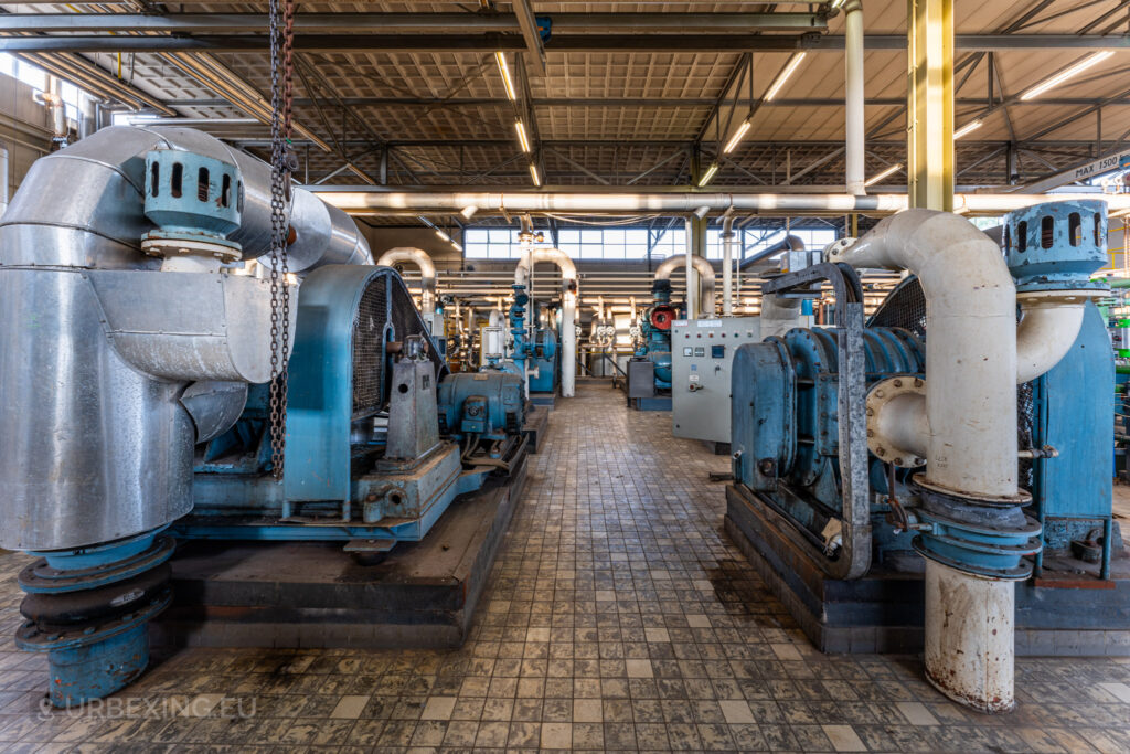 A section inside the abandoned EMGO factory in Lommel, Belgium, featuring large blue industrial machines, blowers, and compressors, connected by an extensive network of pipes, showcasing the decayed industrial infrastructure.