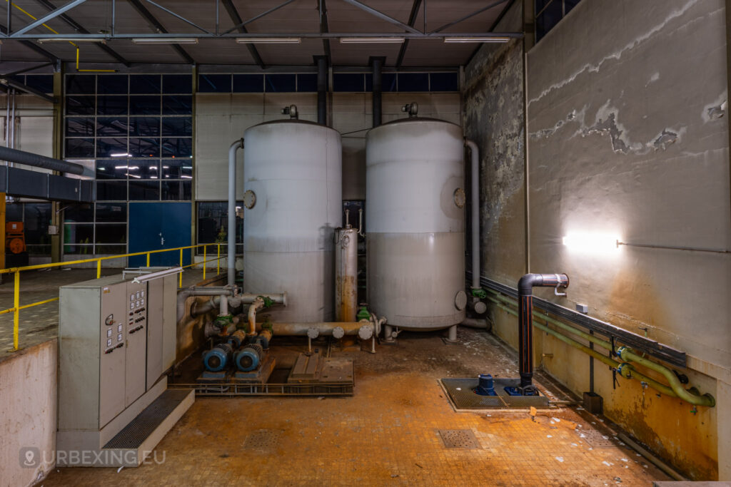 An interior view of an abandoned industrial facility showcasing two large white water tanks. The tanks are connected by a network of rusty pipes and control panels, with peeling paint on the walls and debris scattered across the floor. The dim lighting casts shadows, emphasizing the decay and neglect of the space. The image captures the eerie, silent atmosphere of an urban exploration site, perfect for enthusiasts interested in discovering forgotten industrial locations.