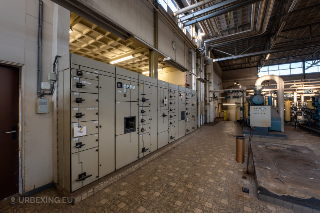 The abandoned EMGO factory in Lommel, Belgium, showcasing a long row of control panels and extensive piping, reflecting the factory's sophisticated water treatment system amidst its current state of decay.