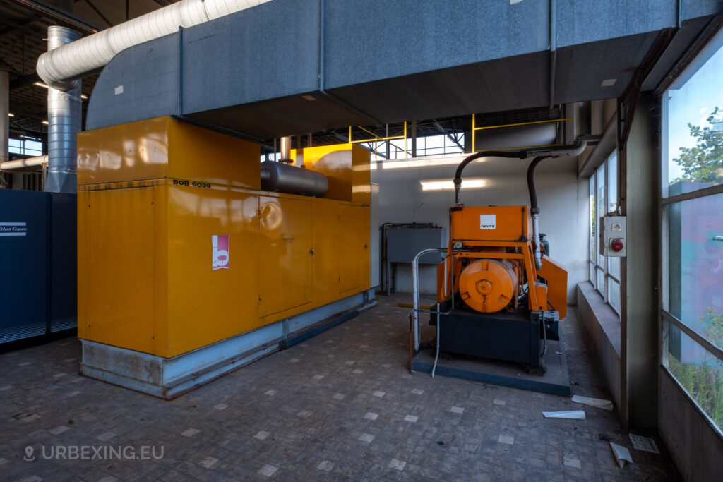 The abandoned EMGO factory in Lommel, Belgium, featuring a yellow diesel generator and a smaller orange Deutz unit, highlighting the factory's extensive power infrastructure amidst its decay.