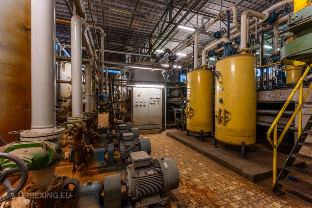 A detailed view inside the abandoned EMGO factory in Lommel, Belgium, showcasing yellow tanks, electric motors, and a complex network of pipes and valves, reflecting the extensive water treatment setup amidst the factory's decayed state.