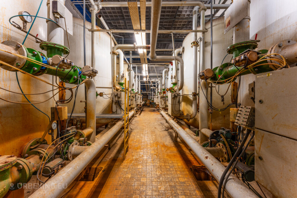 A narrow corridor inside the abandoned EMGO factory in Lommel, Belgium, lined with large tanks and a network of pipes and valves, showcasing the complex industrial infrastructure amidst the decayed state of the facility.