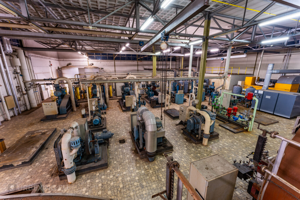 An overhead view of the abandoned EMGO factory in Lommel, Belgium, filled with rows of blue industrial machines and a complex network of pipes, highlighting the extensive and decayed industrial setup.