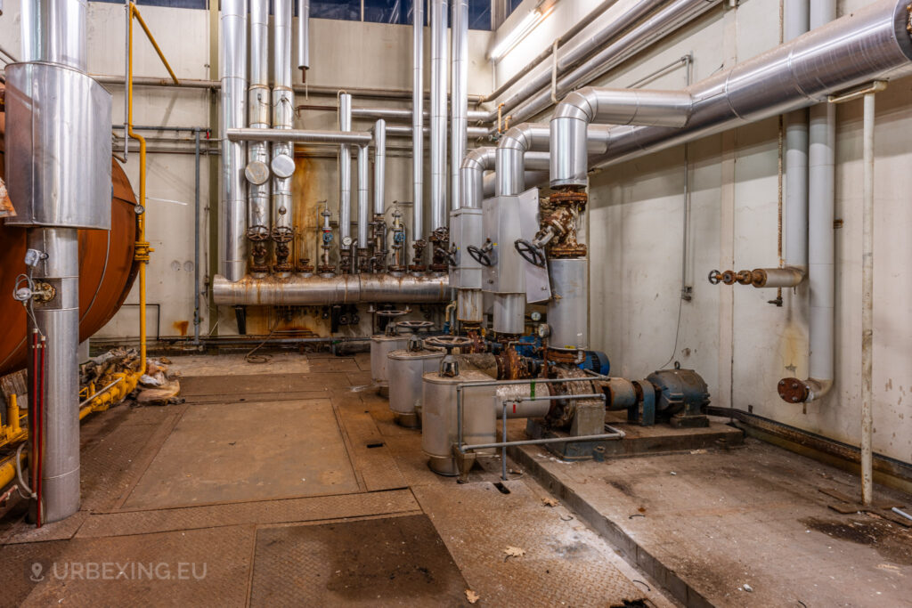 An industrial area inside the abandoned EMGO factory in Lommel, Belgium, showcasing a complex network of metallic pipes, pumps, and valves, highlighting the extensive infrastructure amidst the factory's state of decay.