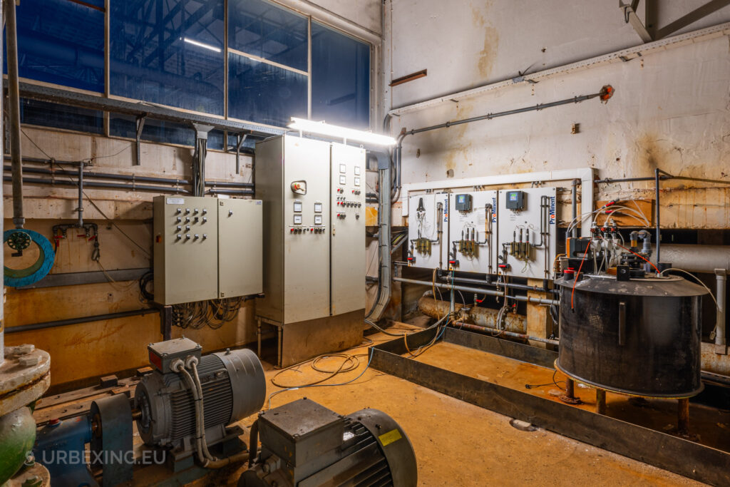 A corner of the abandoned EMGO factory in Lommel, Belgium, featuring control panels, electric motors, and mechanical components, showcasing the decayed state of the factory's water treatment or electrical system.