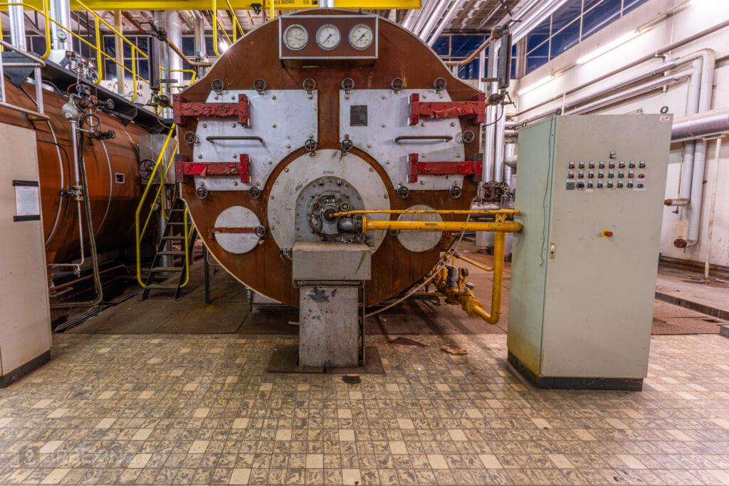 A close-up of an industrial boiler in the abandoned EMGO factory in Lommel, Belgium, showcasing the intricate gauges, pipelines, and control panels amidst the decayed industrial environment.