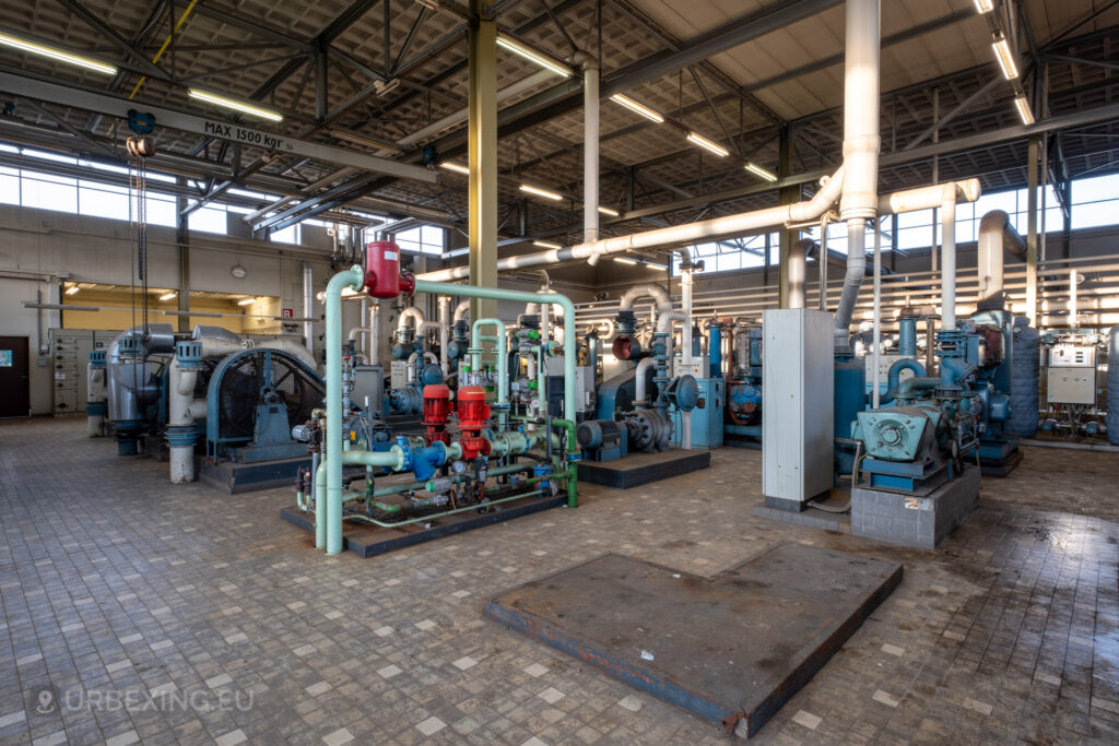 A spacious room inside the abandoned EMGO factory in Lommel, Belgium, featuring an array of water treatment machinery and pipes, highlighting the extensive industrial setup amidst the factory's state of decay.