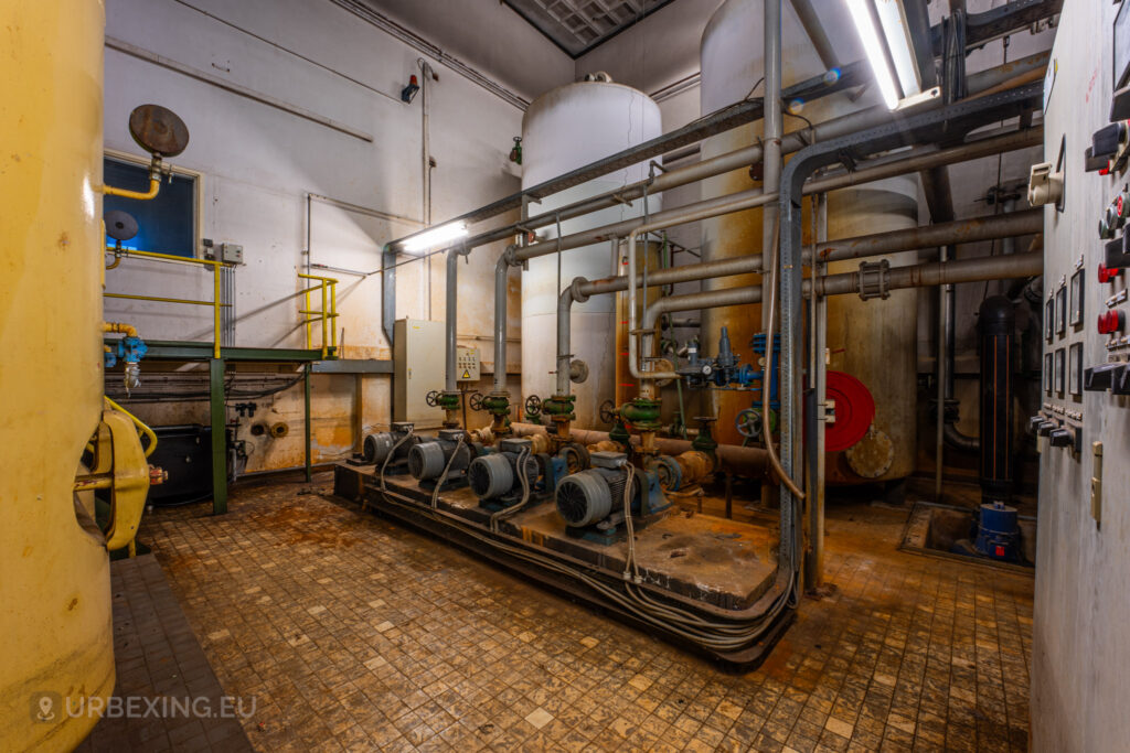 An intricate view inside the abandoned EMGO factory in Lommel, Belgium, showcasing large tanks, electric motors, and an extensive network of pipes and valves, highlighting the decayed state of this former water treatment room.