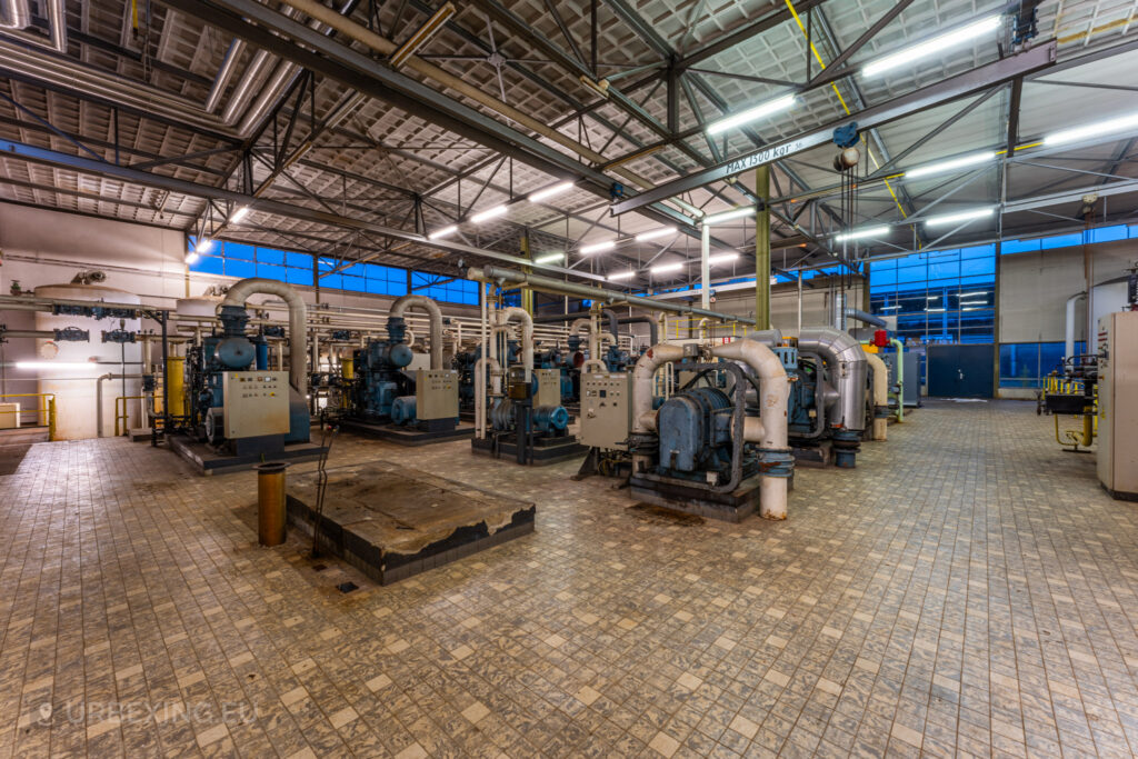 A large room in the abandoned EMGO factory in Lommel, Belgium, filled with blue water treatment machinery, pipes, and control panels, showcasing the extensive industrial setup amidst the factory's state of decay.