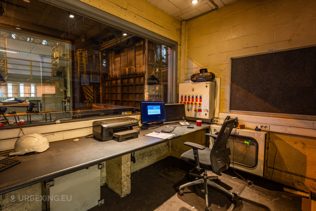 An abandoned control room at the Cockerill Forges & Ringmill factory, featuring a desk with a computer, printer, and control switches. The workspace includes a chair and various items left behind, like a safety helmet and a red cup. The large window provides a view into the adjacent factory floor, showing large industrial machinery and yellow railings. The overall atmosphere is one of disuse and abandonment, with a dusty desk and dim lighting.