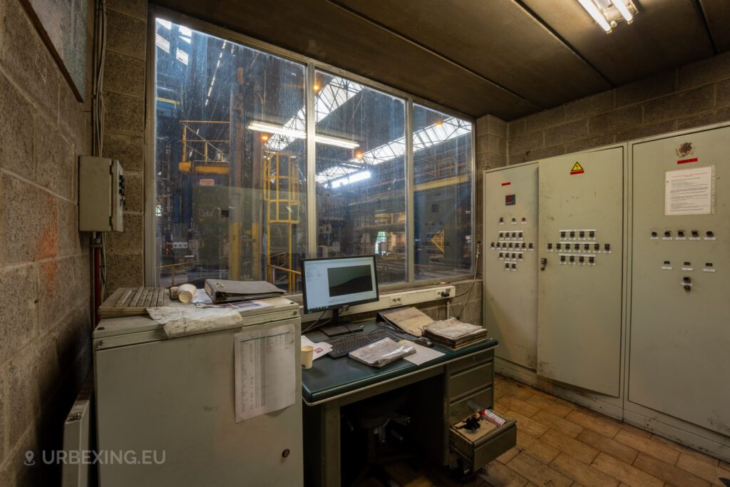 An abandoned control room in the Cockerill Forges & Ringmill factory, featuring a desk with a computer, binders, and scattered paperwork. The room has a large window looking out onto the factory floor, giving a view of the yellow industrial railings and machinery outside. Control panels with numerous switches are mounted against the wall, suggesting the operational role of the room in managing factory processes. Dust and disorganization highlight the long-neglected state of this workspace.