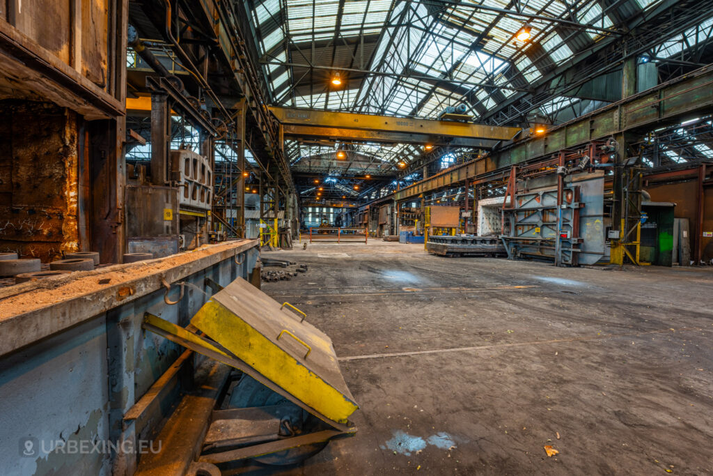 A view of the abandoned Cockerill Forges & Ringmill factory floor, with a large, open furnace and other industrial equipment visible. The scene shows the rusted and weathered state of the machinery, with a yellow safety barrier in the foreground. Overhead, an industrial crane system runs the length of the ceiling, and the expansive floor area is cluttered with grime and scattered remnants of past production. The high ceiling structure, crisscrossed with steel beams, conveys the vastness of the factory space.