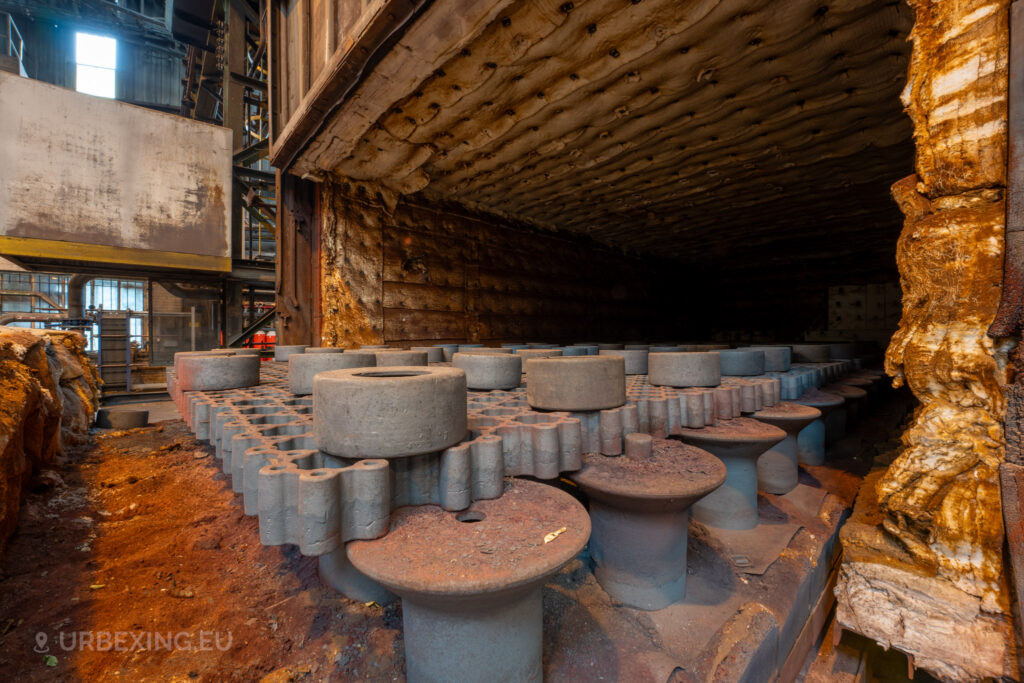 A close-up view inside an abandoned furnace at the Cockerill Forges & Ringmill factory. The interior is lined with rusted, insulated walls, and filled with numerous heavy steel molds resting on a grid-like platform. The molds, covered in rust and dust, are arranged neatly inside the furnace, which shows signs of wear and decay. The image highlights the immense heat-resisting materials of the furnace walls and the machinery that has remained untouched for years.