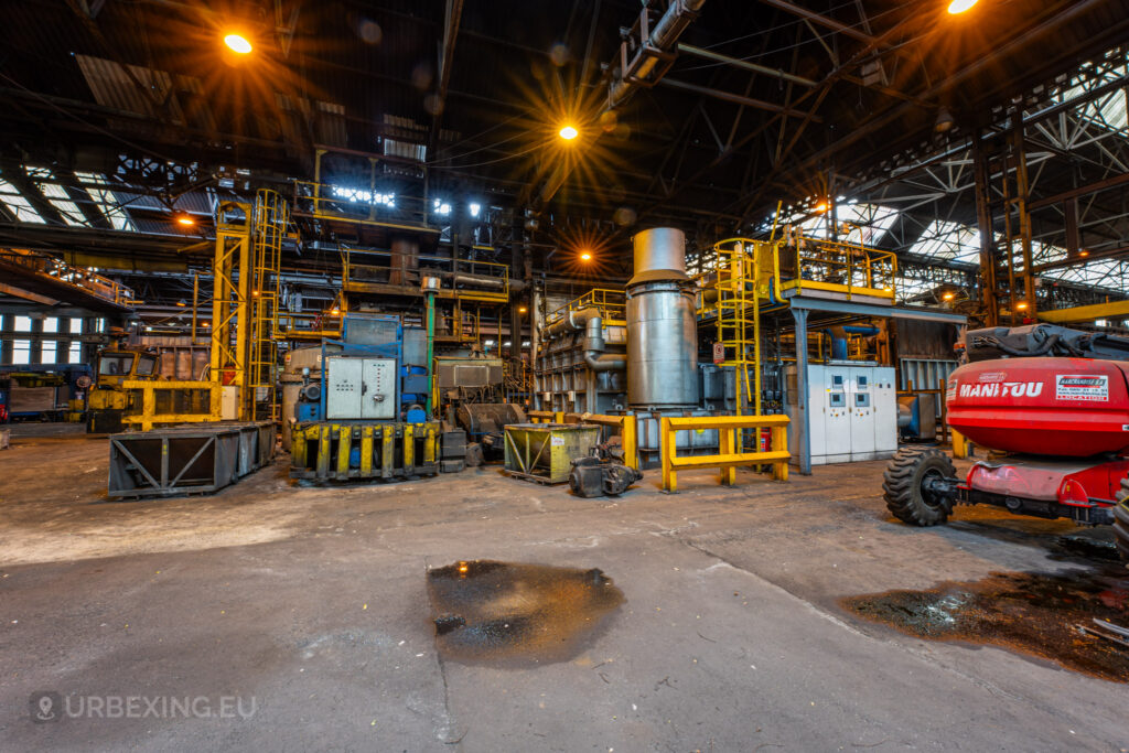 An abandoned industrial processing area inside the Cockerill Forges & Ringmill factory, featuring a collection of large machinery including metal tanks, pipes, and control panels. The factory floor is cluttered, with a yellow safety barrier and a red utility vehicle in view. Overhead, steel beams support a roof with partially broken skylights, letting in light that highlights the industrial decay. The puddles of water and rust-stained surfaces indicate long-term abandonment of this once-busy space.