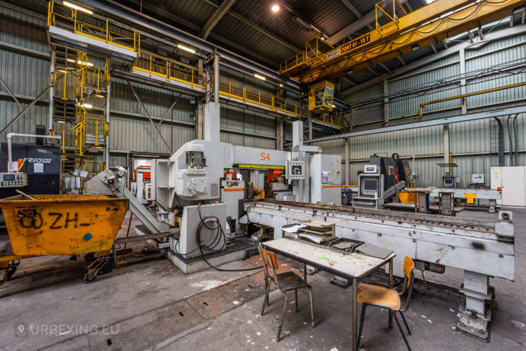 A view of an abandoned manufacturing station in the Cockerill Forges & Ringmill factory, featuring large white and gray industrial machinery marked ‘S4.’ A yellow bin is positioned next to the machine, and a table with scattered papers and two chairs sits in the foreground, suggesting a hastily abandoned workspace. Overhead, a bright yellow crane and a metal walkway are visible, adding to the industrial atmosphere of the high-ceilinged workshop.