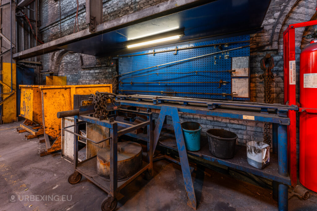 An abandoned workbench inside the Cockerill Forges & Ringmill factory, featuring a blue pegboard with tools, various storage bins, and rusted chains. Yellow metal bins labeled ‘Bois’ and other containers are scattered around, suggesting an organized workspace that has since been left to decay. The fluorescent light above the workbench casts a dim glow on the aged and worn equipment, with rust and grime covering the surfaces, highlighting the state of disuse