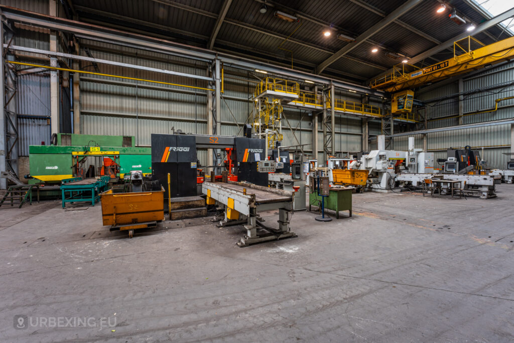 An expansive view of an abandoned workshop inside Cockerill Forges & Ringmill, featuring a variety of heavy machinery and industrial equipment. The machines are painted in bright colors such as green, yellow, and black, and are accompanied by rusting metal bins scattered across the floor. A yellow overhead crane system is visible in the background, suspended from the ceiling, and the workshop space appears vast and empty, with signs of disuse and grime covering surfaces.