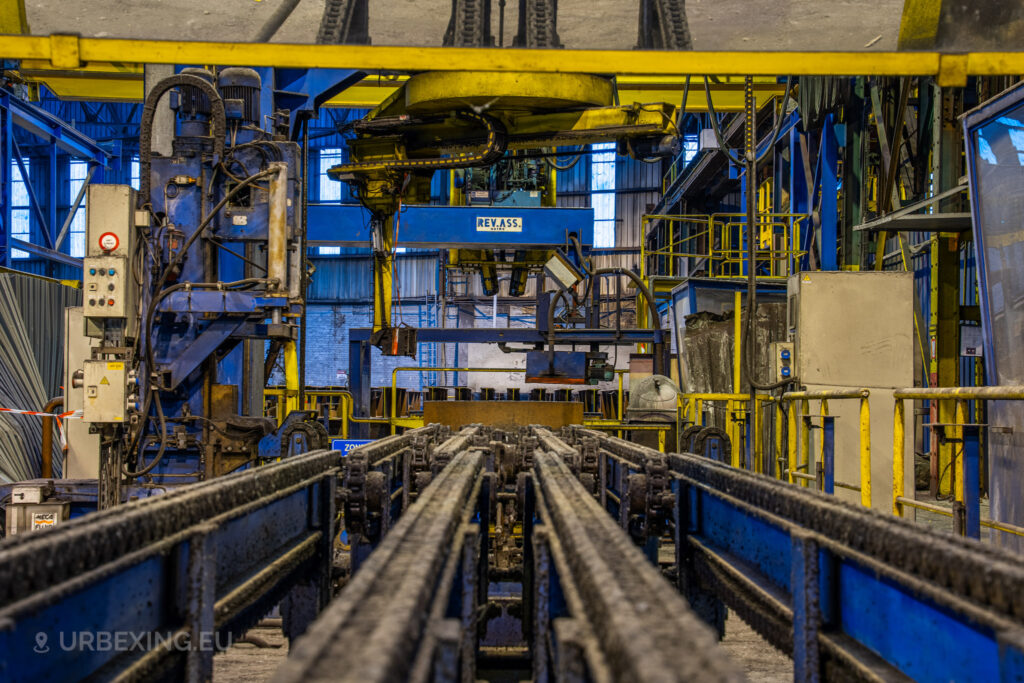 A close-up view of a chain conveyor system in the abandoned Cockerill Forges & Ringmill factory, with overhead machinery labeled ‘REV.ASS’. The chains are rusty, and the surrounding machinery, painted in blue and yellow, shows signs of corrosion and neglect. The background reveals multiple platforms, control panels, and railings that were once part of an extensive steel processing operation. The industrial setting is characterized by its heavy-duty equipment and state of disrepair, conveying an atmosphere of abandonment.