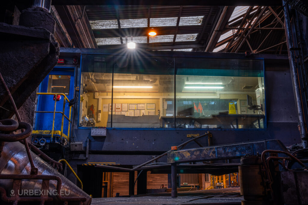 A view of the control room in the Cockerill Forges & Ringmill factory, elevated above the workshop floor. The room, encased in large windows, is partially illuminated, revealing desks, paperwork, and safety gear. A blue door and yellow railing lead up to the entrance. The ceiling above features exposed beams, and a bright light fixture casts a glow across the scene, emphasizing the contrast between the illuminated interior and the dark, industrial surroundings below.