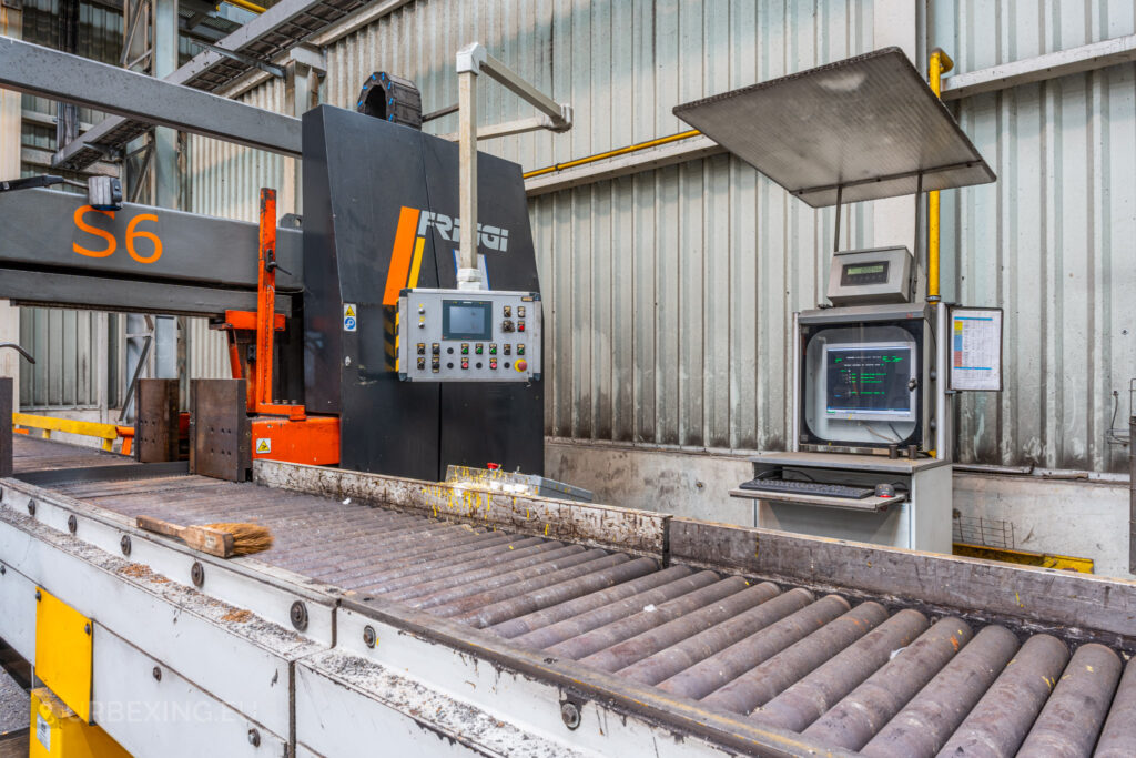 A close-up view of a conveyor system and a control panel at the Cockerill Forges & Ringmill factory. The machine, labeled ‘S6,’ includes a touchscreen interface, various buttons, and warning labels. Next to it, a computer terminal with a display monitor is protected by a metal cover, suggesting its use in managing the conveyor process. The scene depicts an abandoned industrial setup, with a brush left on the rollers and visible signs of grime and wear throughout the area.