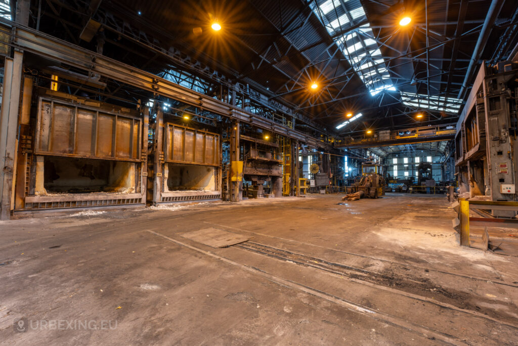 A wide view of the abandoned Cockerill Forges & Ringmill factory floor featuring multiple large, open furnaces with exposed insulation. The scene is illuminated by overhead lights casting a warm glow, revealing the aged and rusted state of the heavy machinery. The expansive floor is marked with old rail tracks leading deeper into the mill, and an industrial vehicle is visible in the distance, adding to the atmosphere of decay and abandonment.