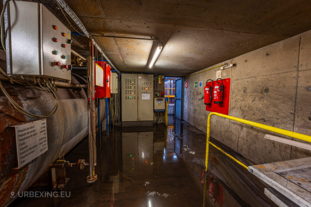 A flooded basement control room in the abandoned Cockerill Forges & Ringmill factory, with control panels, fire extinguishers, and industrial piping lining the concrete walls. The floor is covered in standing water, reflecting the fluorescent lights above and creating a damp, decaying atmosphere. Electrical panels and warning signage are visible, indicating the room’s previous function as an operational area for monitoring factory systems. The entrance door is slightly open, revealing an industrial setting beyond.