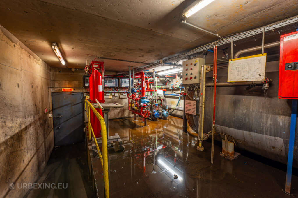 A flooded control room in the abandoned Cockerill Forges & Ringmill factory, with large red valves and control machinery. Water covers the concrete floor, reflecting the overhead fluorescent lights, while various pipes and control panels line the walls. The room is narrow, with industrial equipment showing signs of corrosion and disuse. A heavy metal door and a yellow safety railing add to the industrial feel of this damp, forgotten section of the facility.