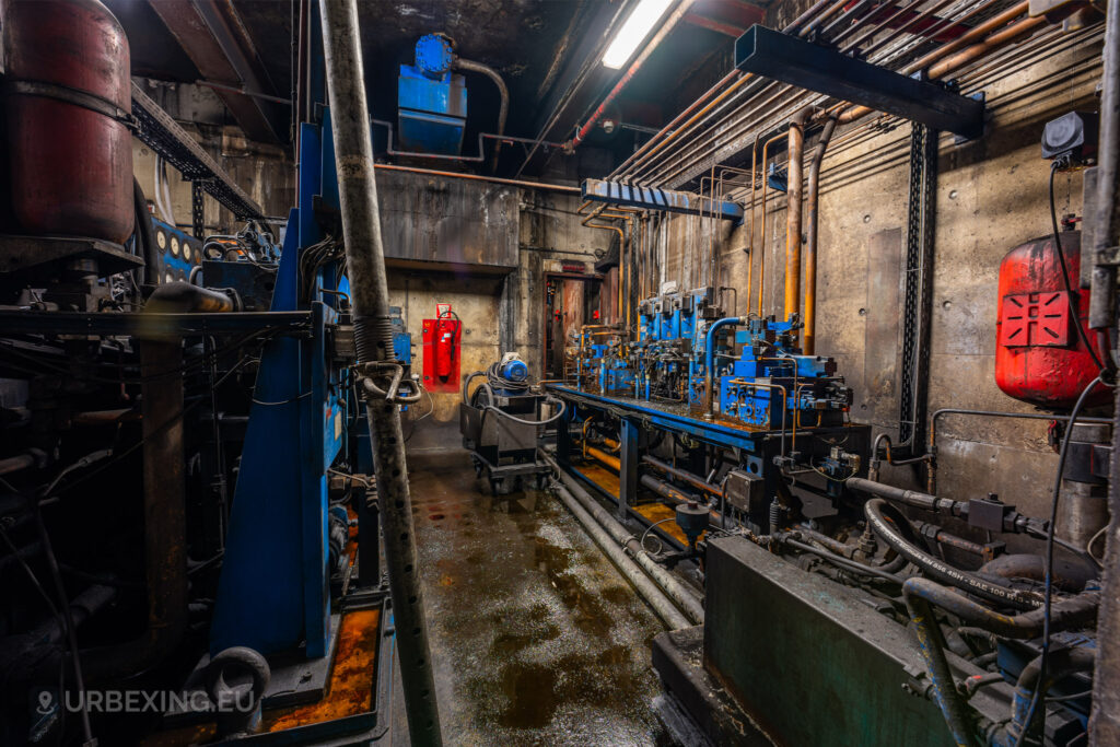 A narrow hydraulic control room in the abandoned Cockerill Forges & Ringmill factory, featuring complex blue machinery, rusted tanks, and a maze of pipes running along the walls. The floor is flooded, and grime covers the equipment, reflecting a state of disrepair. A red fire extinguisher and pressure gauges can be seen on the wall, indicating the once-critical nature of the operations here. The dim lighting and wet surfaces create a gloomy atmosphere within this forgotten industrial space.