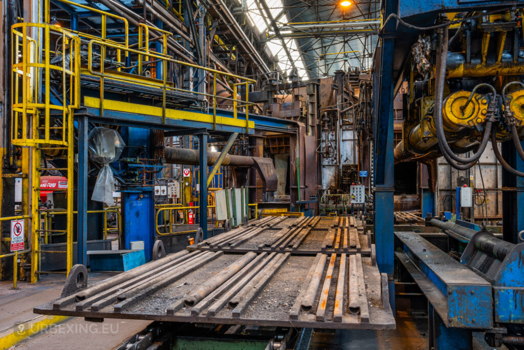 A view of a heavy metal conveyor system in the abandoned Cockerill Forges & Ringmill factory, surrounded by hydraulic machinery and pipes. The conveyor is covered in dust and rusted metal components, indicating years of neglect. Yellow railings and platforms are visible, as well as various blue and yellow machinery parts, showing the once intricate process flow of the steel mill. The overhead ceiling features steel beams and industrial lighting, adding to the complex and rugged atmosphere of the mill.