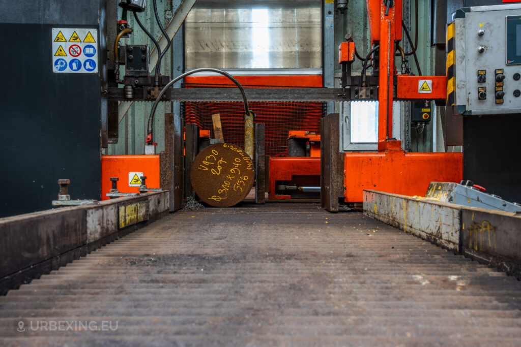 A close-up view of a large cylindrical metal rod positioned in a cutting machine at the abandoned Cockerill Forges & Ringmill factory. The orange and black heavy-duty equipment surrounds the metal piece, which has markings written on its surface. The conveyor mechanism leading to the machine and safety symbols are visible, indicating the industrial nature of the process. The scene captures a moment frozen in time in a disused steel production environment.