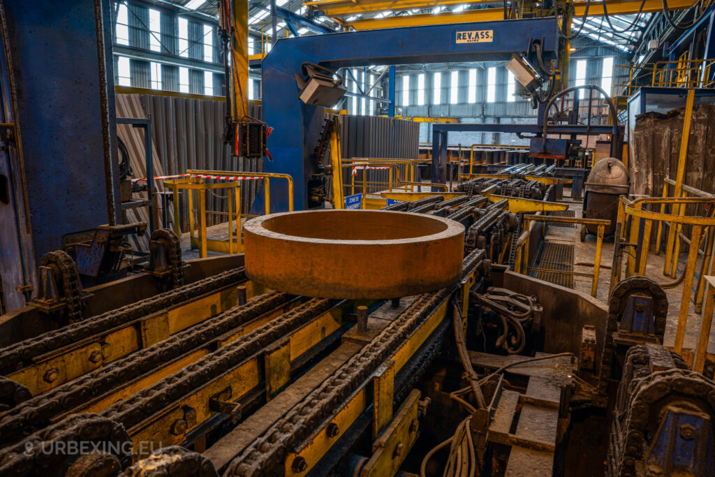 A large metal ring sits on a chain conveyor in the abandoned Cockerill Forges & Ringmill factory. The blue and yellow machinery around it is covered in dust and grime, indicating long-term disuse. Overhead, a blue mechanical arm labeled ‘REV.ASS’ is positioned above the machinery. The factory space is filled with yellow safety railings and industrial equipment, with light filtering through the high windows, highlighting the aged and rusted state of the facility.