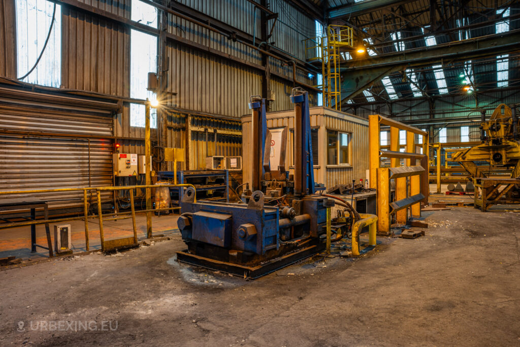 A hydraulic press unit sits abandoned in the Cockerill Forges & Ringmill factory, painted in blue and surrounded by other industrial structures. The press is positioned near a small control office building with a window, and yellow safety barriers mark off different work zones. The ceiling above is a steel frame structure with skylights, adding to the worn-out industrial ambiance of the workshop. The floor is covered in grime, reflecting years of disuse in this deserted industrial setting.