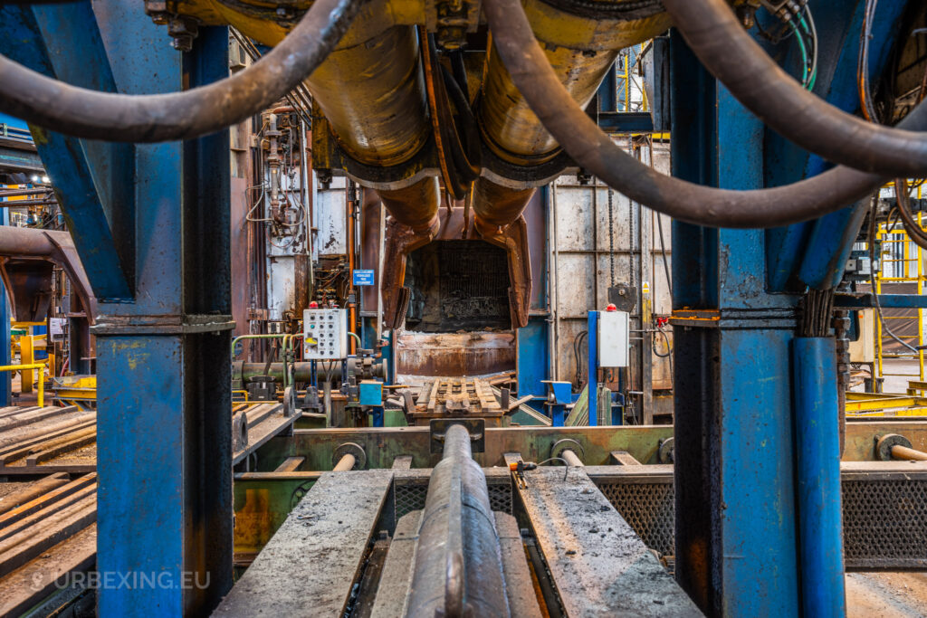 A close-up view of a large hydraulic press and conveyor system inside the abandoned Cockerill Forges & Ringmill factory. Massive hydraulic cylinders dominate the foreground, with cables and pipes running along their length. The background reveals part of a furnace opening and various metal frameworks covered in rust and grime, indicating the years of disuse. Control panels and industrial equipment are scattered around, capturing the heavy-duty machinery and complex workings of the steel mill.