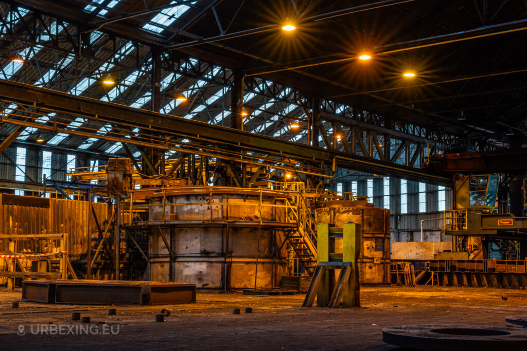 A view of a massive industrial furnace at the Cockerill Forges & Ringmill factory, surrounded by steel beams and heavy machinery. The rusted structure is highlighted by warm, orange lighting, and stairs lead up to a platform for maintenance access. Overhead, large beams and supporting structures dominate the high ceiling, and a yellow glow from the lights adds to the industrial atmosphere. The scene captures the sheer scale and complexity of the furnace, now idle in the abandoned steel mill.