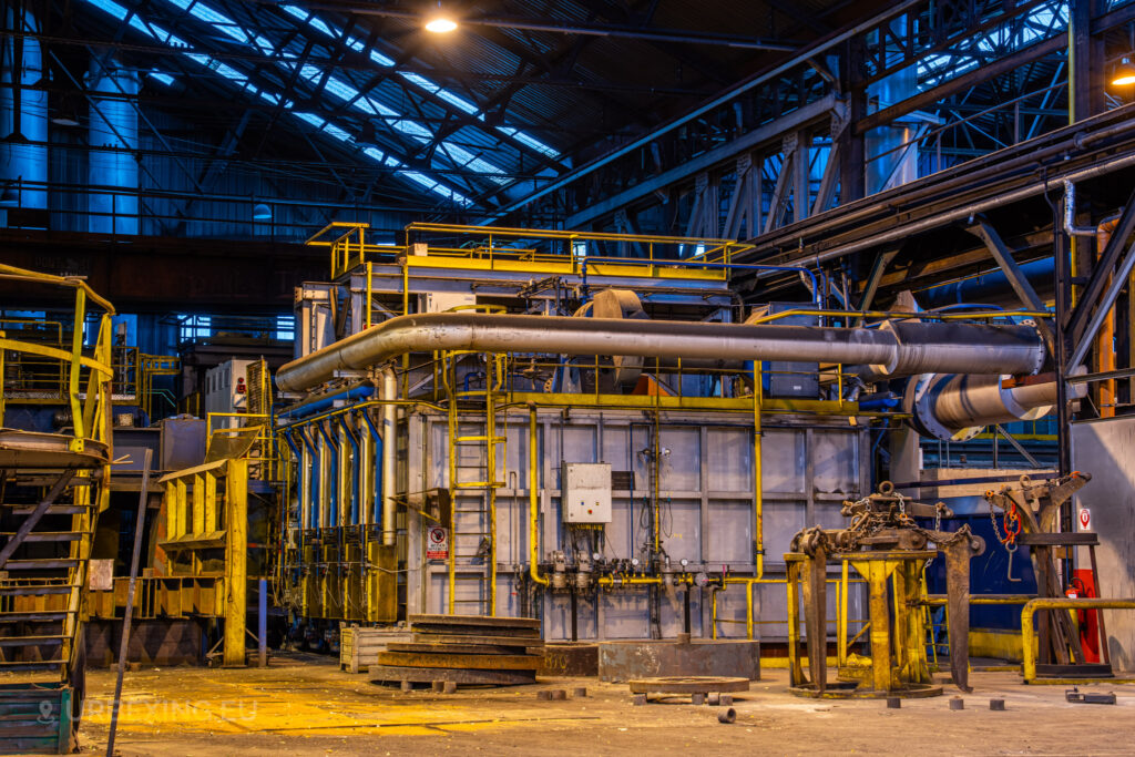 A large industrial furnace surrounded by a complex network of piping and metalwork in the abandoned Cockerill Forges & Ringmill factory. The machinery is framed by yellow railings and platforms, with heavy pipes running across the structure, indicating the scale of the steel processing operations. Rust and grime cover much of the equipment, while dim lighting illuminates the scene, creating a stark industrial atmosphere. The open space and exposed steel beams above add to the vastness of the facility.