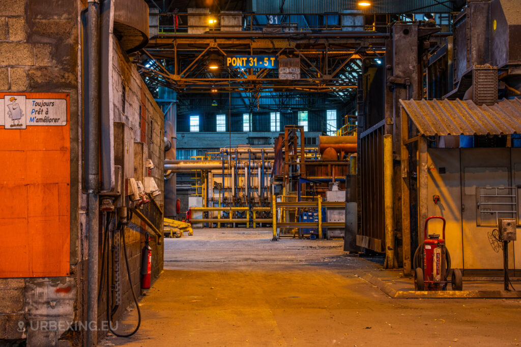 A view down a hallway in the abandoned Cockerill Forges & Ringmill factory, showing industrial piping and machinery in the background. The scene is framed by walls of exposed brick and metal, with a sign in French that reads ‘Toujours Prêt à M’améliorer,’ which translates to ‘Always Ready to Improve.’ Overhead, a large crane marked ‘PONT 31 - 5 T’ spans the ceiling, while a fire extinguisher and various utility equipment are seen on the floor, emphasizing the aged and functional atmosphere of the former steel mill.