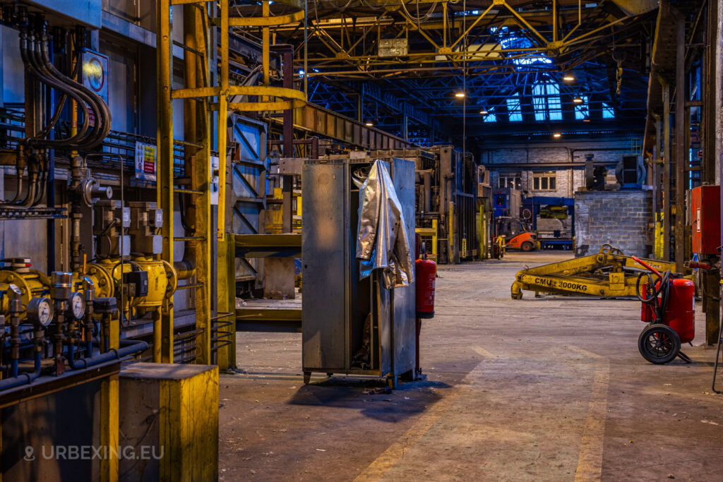 A view down an industrial hallway in the abandoned Cockerill Forges & Ringmill factory. The scene features pipes, yellow machinery, and control systems lining the wall. In the middle, a metallic jacket hangs from an open cabinet, giving a sense of abandonment. Fire extinguishers and a hydraulic lift labeled ‘CMU 3000KG’ are scattered throughout the area. The dim lighting and exposed metal structures create a gritty industrial atmosphere, emphasizing the unused state of the equipment.