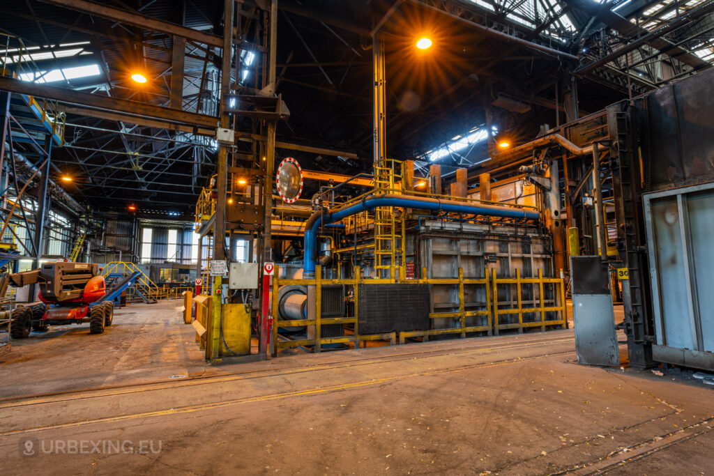 An interior view of the abandoned Cockerill Forges & Ringmill factory, showcasing heavy industrial machinery and vibrant blue piping. The structure is surrounded by yellow railings, and safety signage is visible. The overhead ceiling is supported by steel beams, with lights casting a warm orange glow across the space. A red utility vehicle is parked nearby, and the scene conveys the industrial complexity and the current state of abandonment of this steel processing facility.