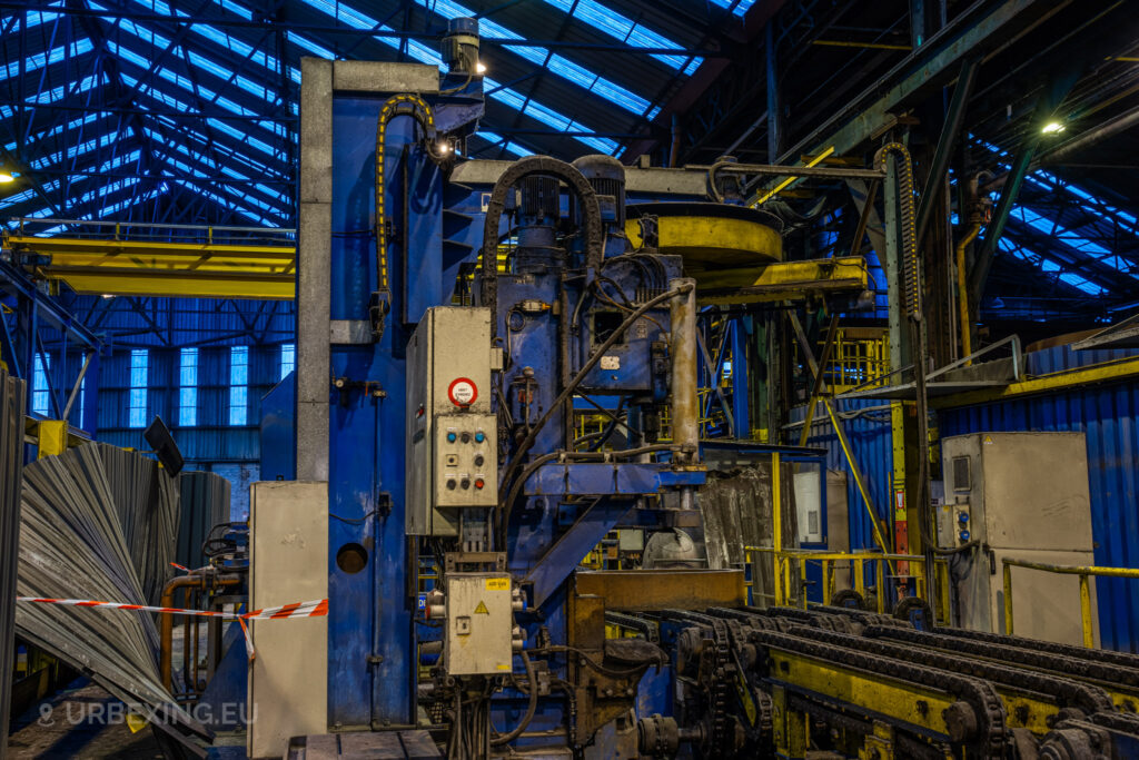 A close-up of industrial machinery at the abandoned Cockerill Forges & Ringmill factory, featuring a large blue and yellow machine with various control boxes and electrical components. The machine appears heavily worn, with rust and grime accumulated on the surfaces. The conveyor system in the foreground is part of the production line, while overhead steel beams and translucent roof panels filter blue-toned natural light into the scene, creating an industrial yet somber atmosphere.