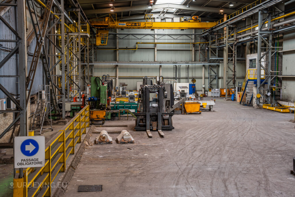 A wide view of an abandoned industrial workshop at Cockerill Forges & Ringmill, showcasing machinery, a forklift, and an overhead crane system. The space is filled with various pieces of equipment, including a large green cutting machine and several orange bins. A yellow barrier and a ‘Passage Obligatoire’ sign indicate organized work zones. The walls are lined with metal frames and walkways, while overhead cranes extend across the ceiling, providing a sense of scale and complexity of this former manufacturing hub.