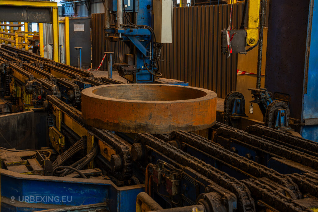 A close-up view of a large, rusted metal ring sitting on a chain conveyor system at the abandoned Cockerill Forges & Ringmill factory. The machinery, painted in yellow and blue, shows significant rust and wear, indicating years of disuse. The conveyor system is surrounded by yellow safety railings, and industrial control boxes and cables can be seen around the area. The scene highlights the heavy-duty nature of the steel processing operations once carried out here.