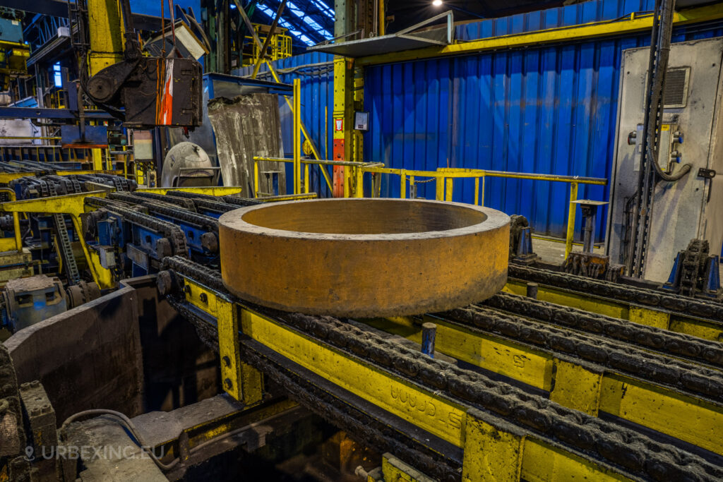 A large metal ring sits on a yellow and blue conveyor system at the abandoned Cockerill Forges & Ringmill factory. The chain-driven conveyor is heavily rusted, indicating long-term disuse. Yellow safety railings and a blue partition wall can be seen in the background, alongside other industrial equipment. The machinery’s worn state, with grime and corrosion covering the components, reflects the overall abandoned condition of this once-functioning steel processing facility.