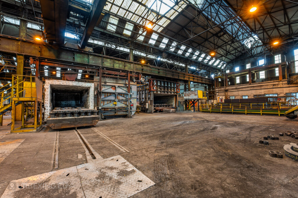 An open furnace in the abandoned Cockerill Forges & Ringmill factory, showing exposed insulation and a rail track leading into its interior. The scene captures the vast factory floor, with numerous pieces of heavy industrial equipment and structural beams overhead. Yellow safety railings and platforms surround the area, and natural light filters in through the skylights above. The image highlights the expansive scale of the steel mill and the disused state of its once-operational machinery.