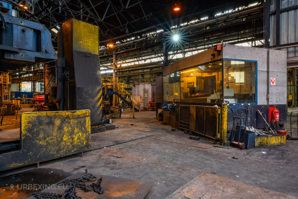 A view of an operator cabin inside the abandoned Cockerill Forges & Ringmill factory, surrounded by large industrial machinery. The cabin features large glass windows, through which control equipment is visible. Yellow safety barriers and old chains are scattered around the dusty concrete floor, and the high ceiling of the forge hall is lined with steel beams. Overhead lights create a dim, industrial atmosphere, giving the sense of a once-active heavy manufacturing environment now left to decay.